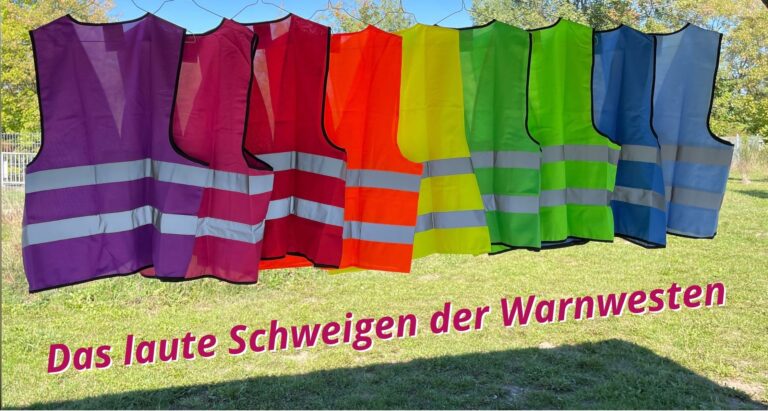 Neun Warnwesten in unterschiedlichen Farben hängen nebeneinander im Freien mit dem Schriftzug: Das laute Schweigen der Warnwesten